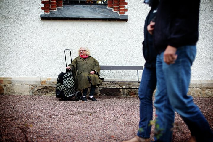 Eldre kvinne sitter på benk mens folk går forbi.