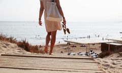 Ryggen til en kvinne som går ned mot stranden med sandaler i hånden.