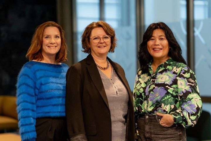 Tre profesjonelle kvinner står smilende side om side og poserer for et bilde. Bakgrunnen antyder et moderne kontorlandskap.